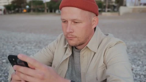 Stylishly dressed young man is sitting on the beach and using the phone Video stock 222603207