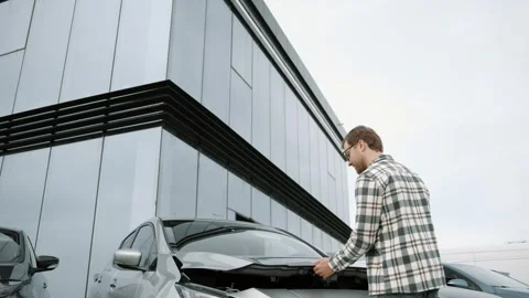 Successful male customer looking inside opened car hood of silver electric car Stock Footage 241417898