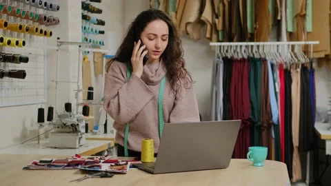 Successful tailor entrepreneur is talking on mobile phone and using laptop. Woma Stock Footage 151385355