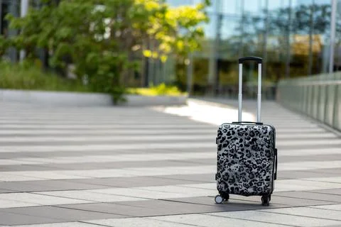 A suitcase stands alone on a stone path outside a modern building during da.. Stock Photos
