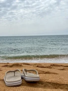 Summer beach scene with flip flops on sand, ocean waves, and cloudy sky conce Stock Photos