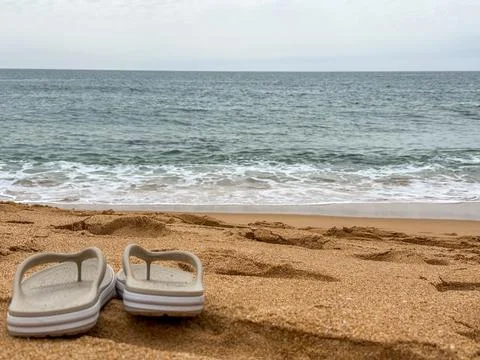 Summer concept. flip flops on sandy beach by ocean waves under cloudy sky. Stock Photos