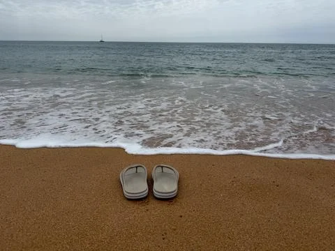Summer concept flip flops on sandy beach with ocean waves and a sailboat unde Stock Photos