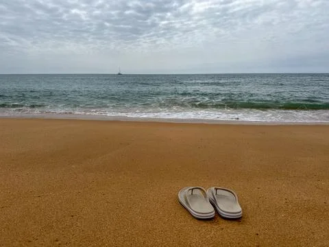 Summer concept: flipflops on a sandy beach with ocean waves, a sailboat, and  Stock Photos
