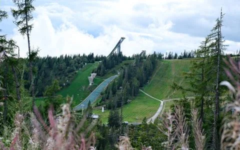 A summer view of a ski jump complex on a green, forested hill, with a cloudy Stock Photos