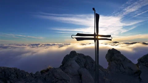 Sun behind religious cross in top of alps mountains peak above foggy clouds Stock Footage 295037833