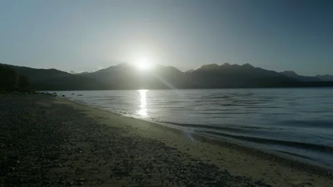 Sun going down behind mountains with waves breaking on lake shore Stock Footage 260369973