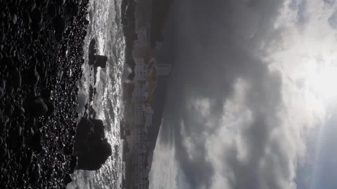 The sun peeking through gray clouds, Waves crash against the black sand shore Stock Footage 266927037