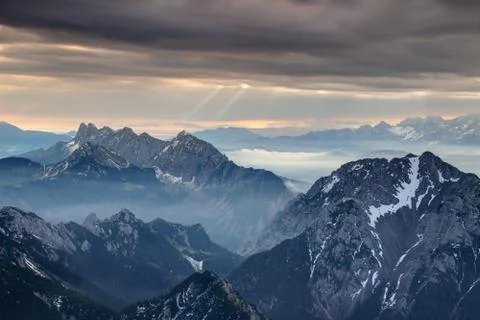 Sun rays burst through dark gray clouds over misty jagged peaks Stock Photos