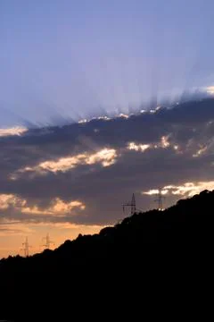 Sun rays shine through clouds with silhouettes of electric poles on the horiz Stock Photos