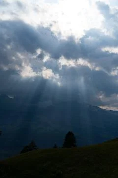 Sun rays shining trough the clouds in the mountains, a hill and trees in fron Stock Photos