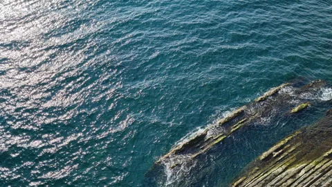 Sun reflecting on a blue water surface near a rocky coast Stock Footage 286012647