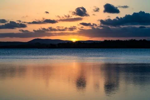 The sun sets behind a mountain, casting warm light across a tranquil lake Stock Photos