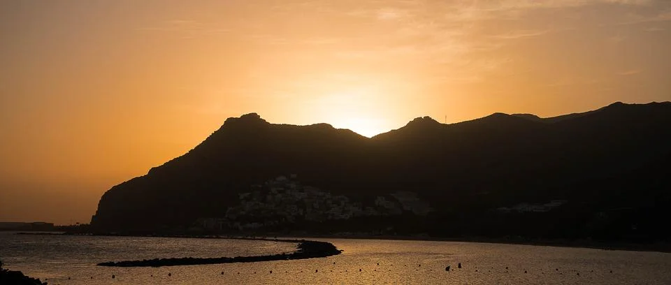 The sun sets behind a mountain range, casting a warm glow over a peaceful bod Stock Photos
