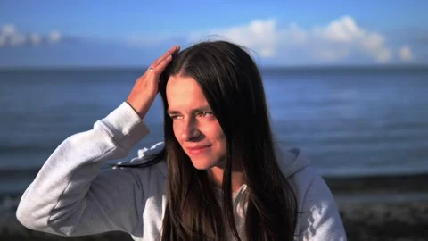 As sun sets behind sea, a young brunette woman stands by shore gently fixing her Stock Footage 282906325