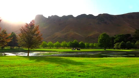 The sun sets over high mountains and illuminates the lake and green meadow Stock Footage 131774169