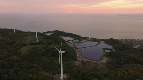 Sun sets over ocean by coastal wind turbines and solar panels producing green Stock Footage 246429154