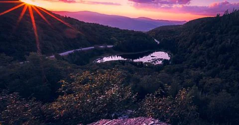 The sun sets over the road cutting through the mountains, by the pond. Stock Photos
