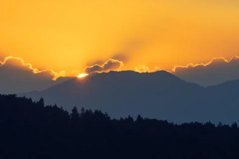 Sun setting behind mountain range and silhouette of spruce forest in foreground Stock Photos