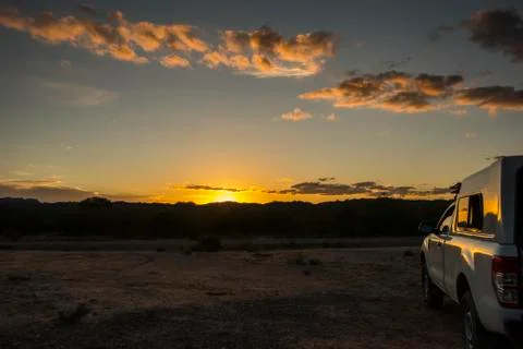 The sun is setting behind a range of hills, lighting up the few clouds in orange Stock Photos