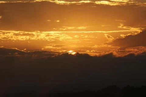 Sun setting behind silhouettes clouds and mountain range with sunlit sky Stock Photos
