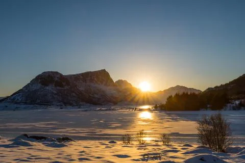 Sun setting behind snow-clad mountains over frozen lake in dreamy winter land Stock Photos