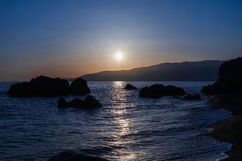 Sun setting over calm sea with silhouette rocky coastline Stock Photos