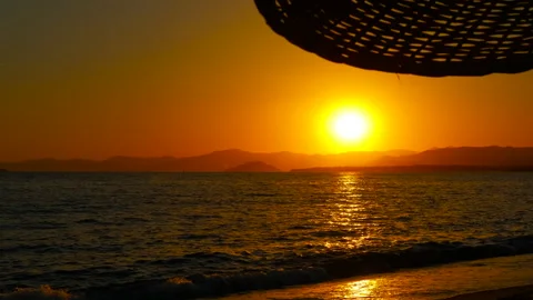 Sun setting over ocean seen from underneath wicker beach umbrella Stock Footage 303756691