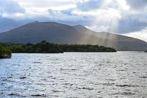 The sun shines through clouds over a lake with mountains Stock Photos