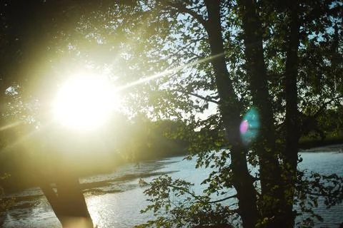 The sun shines through the trees reflecting on the water under sky Stock Photos