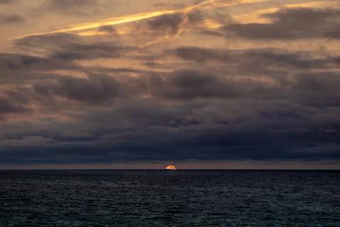 The sun is slowly setting over the vast ocean under a cloudy sky Stock Photos