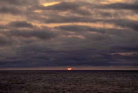 The sun is slowly setting over the vast ocean under a cloudy sky Stock Photos