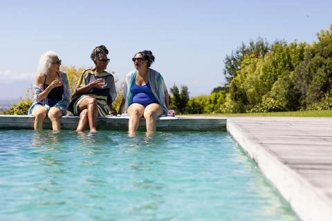 Sun is warming water, causing three women sitting on pool edge dipping feet and Stock Photos