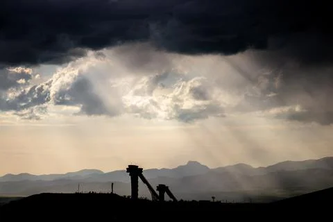 Sunbeams piercing through dark clouds over silhouetted landscape and struct.. Stock Photos