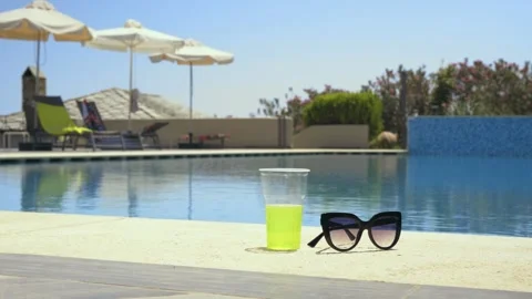 Sunglasses and a drink beside a pool under a bright sunny sky, holiday vibe Stock Footage 328664451
