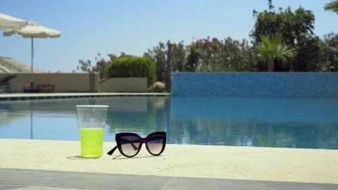 Sunglasses and a drink sit on the edge of a tranquil pool under a clear sky Stock Footage 328658235