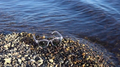 Sunglasses lie on a pebble beach, waves splash at the shore. Stock Footage 89786939