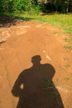 Sunlight casts a shadow of a person on dirt path. Green grass and trees sur.. Stock Photos