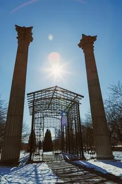 Sunlight comes down through a structure made of columns in a park. Snow lies  Stock Photos