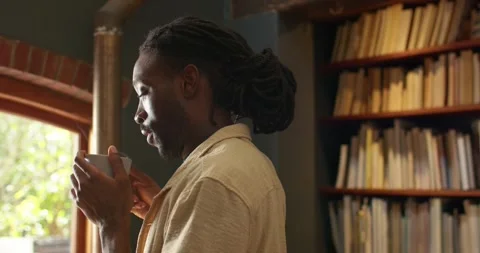 Sunlight drawing eye, African man in shirt in study by arched window sipping Stock Footage 343368194