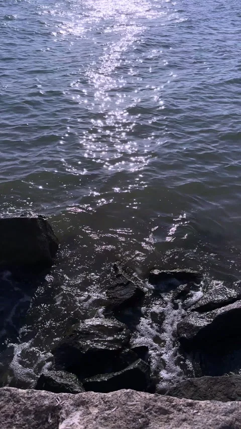 Sunlight sparkling on the dark blue ocean waves hitting black coastal rocks. Stock Footage 341464484