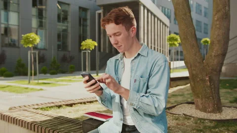 At sunlight student young handsome man with red hair sitting on a bench near Stock Footage 142049944