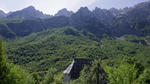Sunlit rustic cabin nestled in lush greenery beneath jagged peaks of Albania's Stock Footage 338426546