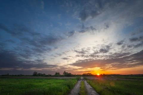 Sunset and evening clouds over meadow with dirt road Stock Photos
