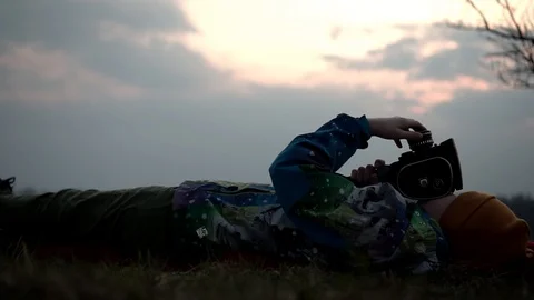 Sunset. The boy is lying on the grass and taking pictures with a movie camera Stock Footage 128330157