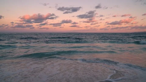 A sunset casts warm colors over a beach with a distant sailboat Stock Footage 312233381