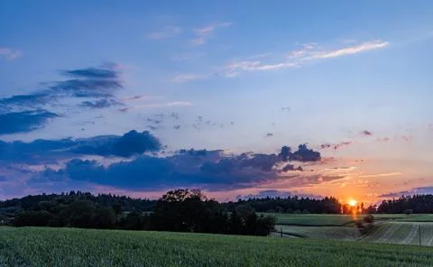 Sunset in a cloudy sky over a hilly rural valley with forests and wheat fields Stock-Fotos