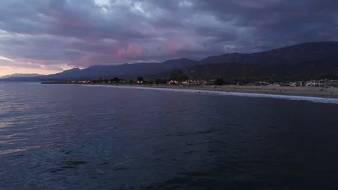 Sunset Drone Towards Beach Over Waves With Homes Mountains and Vibrant Sky 库存影片 100665778