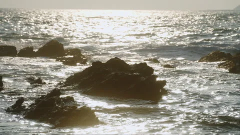 Sunset glistens on ocean as waves crash against rocky shore. Golden hour Stock Footage 272173871