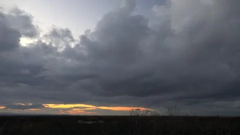 Sunset glowing behind dark nimbus clouds over flat plain landscape under sky Stock Footage 339374057